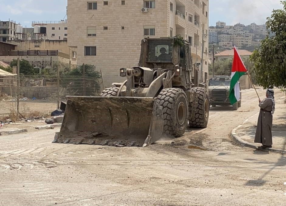 Un hombre ondea una bandera palestina al paso de una excavadora militar que destruye la insfraestructura de la ciudad de Yenín. (Foto: Redes Sociales) 