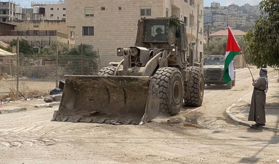 Un hombre ondea una bandera palestina al paso de una excavadora militar que destruye la insfraestructura de la ciudad de Yenín. (Foto: Redes Sociales)