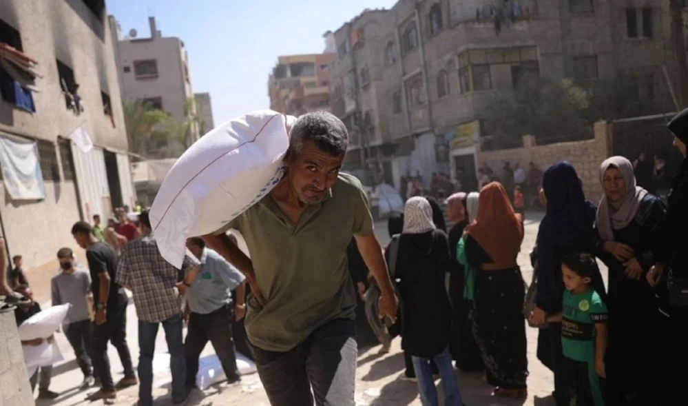Hombre palestino carga un saco de harina, mientras otros esperan en centros de distribución en Gaza.